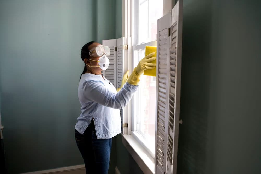 Woman wearing gloves and a mask cleaning a window with a sponge indoors.